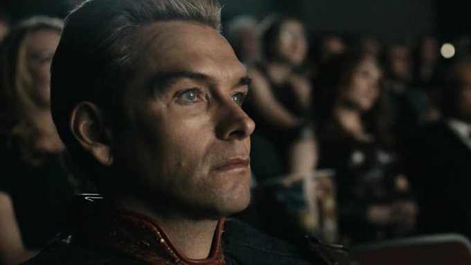 A close-up of a blond man in a dark uniform looking upward with a serious expression, appearing to be in a crowded venue.