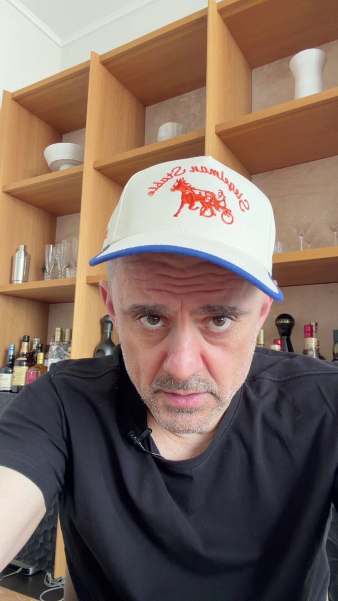 The image features a man wearing a white baseball cap with red lettering, which has "Veefriends. Chrome" written on it. He appears to be posing for a photo or selfie while standing next to a wooden shelf filled with various items such as bottles and wine glasses. The background of the image is blurred, but there are some shelves visible behind him.