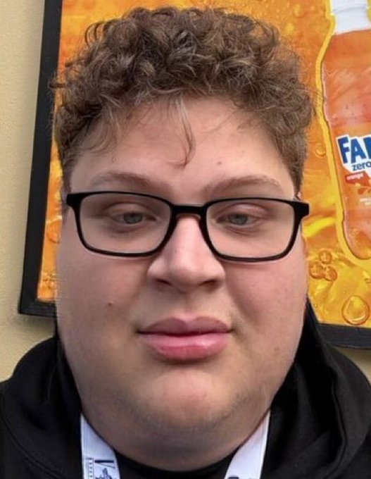 A close-up selfie of a young man with curly brown hair wearing black-framed glasses and a black hoodie, with a Fanta Zero advertisement visible in the background.