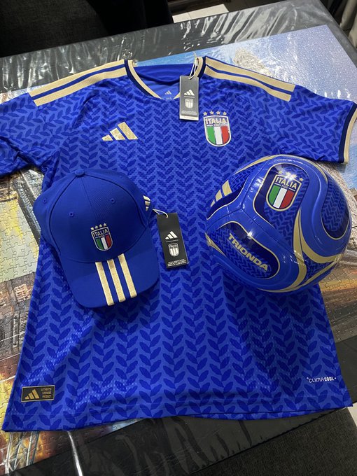 An Italy national team merchandise set including a blue 'ITALIA' Adidas jersey, a matching cap, and a Honda-branded soccer ball, all laid out on a silver surface.