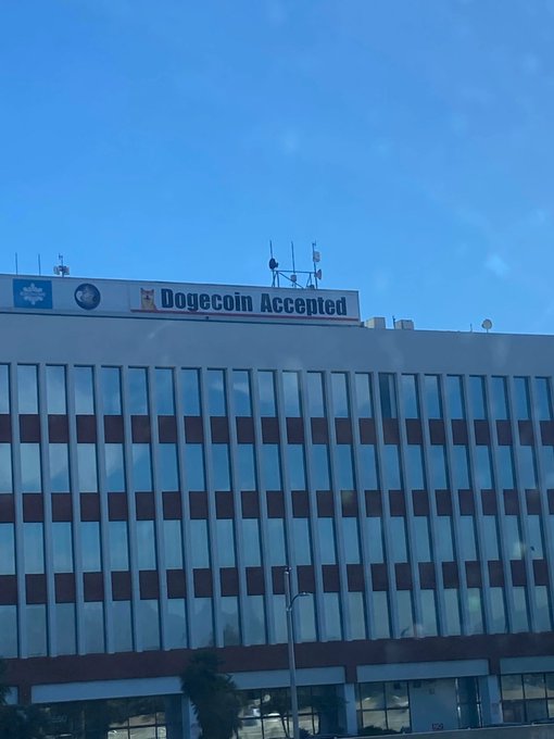 A building rooftop displays a large banner reading 'Dogecoin Accepted' next to a small Doge/Shiba Inu logo, with antennas visible against a clear blue sky.