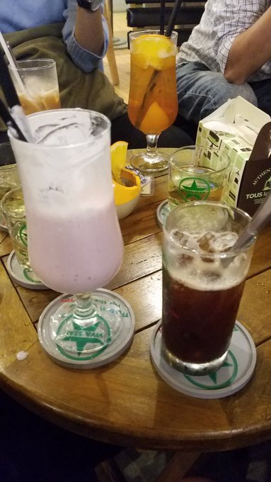 Table at a bar with several drinks including a tall glass of orange juice, a glass of pink milk/yogurt drink on a 'BIERE SAIGON' coaster, and an iced coffee, with people seated around the table.