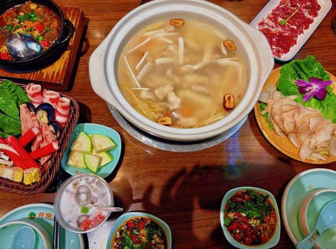 A spread of Asian hot pot dishes including a clear broth pot with mushrooms and meat, raw beef slices, crab sticks, zucchini, dipping sauces, and condiments on a wooden table.