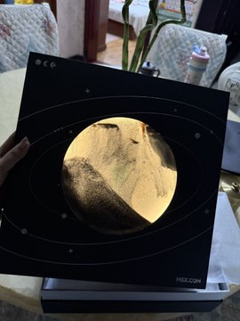 A hand holding a black square package or card featuring a gold metallic circle resembling a planet or moon, with "MBX.COM" printed in the bottom corner.