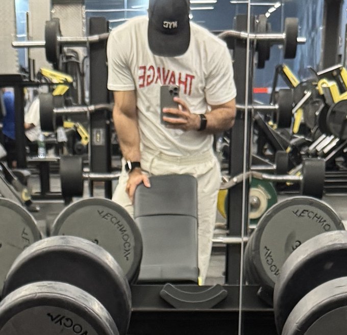 A mirror selfie at the gym showing a man in a "THAVAGE" t-shirt and "NYC" cap taking a photo while leaning on a Technogym bench, surrounded by dumbbells and weight racks—capturing the go-gym grind motivation described in the post.