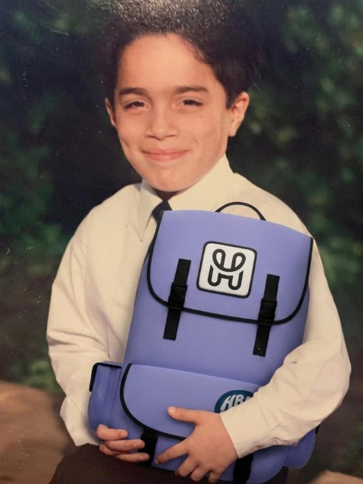A vintage school portrait of a young boy holding a purple backpack featuring a distinctive 'u' logo.