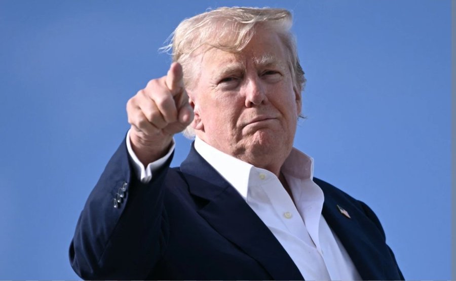 A photograph of Donald Trump in a navy blazer and white shirt, pointing his finger forward against a clear blue sky.