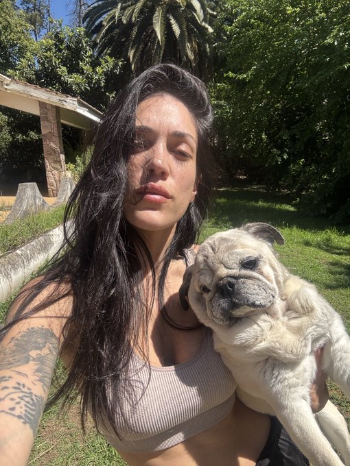 A selfie of a woman with dark hair and tattoos wearing a beige top, holding a small pug in a grassy yard with palm trees in the background.