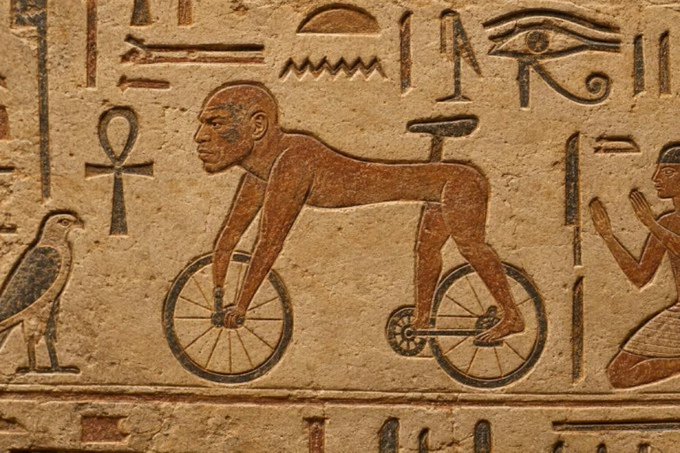 An ancient Egyptian stone relief carving depicting a figure riding a two-wheeled bicycle-like contraption, surrounded by hieroglyphics including ankh symbols and an Eye of Horus.