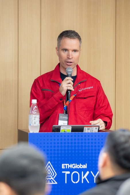 A man in a red 'Curvegrid' jacket speaks into a microphone while standing at a podium labeled 'ETHGlobal TOKYO'.