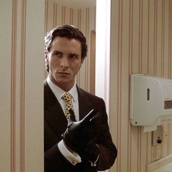 The image features a man dressed in a suit, standing in front of a bathroom mirror with his hands in his pockets. The man appears to be wearing a tie that has leopard print accents. The background shows a bathroom setting with a white paper towel dispenser and a sink visible. The post is related to the movie "@NomismaNetwork" as it features an image of the actor, who is dressed in a suit and holding his hands in his pockets.