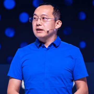 A man wearing glasses and a blue polo shirt, appearing to be giving a speech or presentation on a stage with a dark, blurry background.
