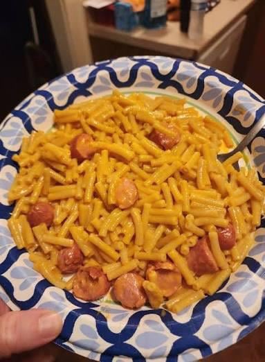 The image features a plate of macaroni and cheese with sausage, which appears to be a delicious meal. The plate has been placed on top of a dining table, ready for someone to enjoy their dinner. In the background, there are two people present - one person is holding the plate up while another person is standing nearby.

The image also contains text that reads "how do I tell my beautiful daughter that she can't play softball this summer? Its because DADDY SPENT ALL HIS MONEY ON MUTANT APES AND BUDLIGHTS IM BACK TO EATING KRAFT DINNER AND HOTDOGS LIKE A BROKE PIECE OF SHIT". This message seems to be a playful way of expressing disappointment in the daughter's desire to play softball, as it is related to the plate of food on the table.
