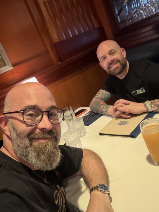 Two smiling bald men pose together at a restaurant table, with a glass of orange-colored beverage visible.