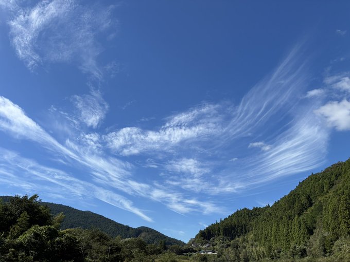 A bright blue sky filled with wispy, streaking cirrus clouds stretches over a valley between two green, forested mountain slopes.