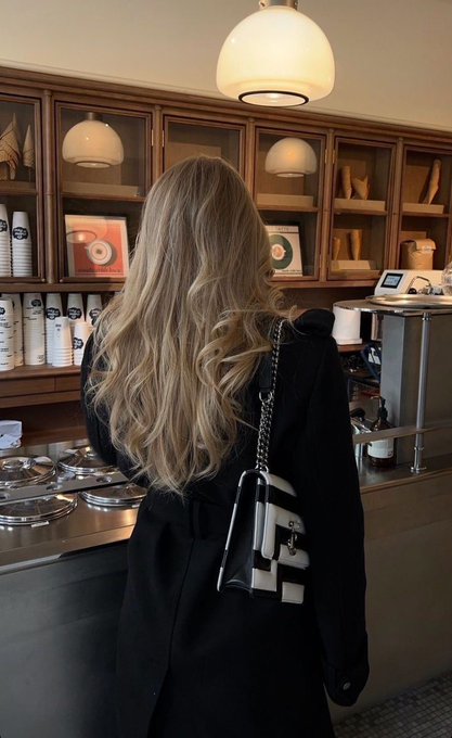 A woman with long, wavy blonde hair, wearing a black coat and carrying a black and white Chanel handbag, stands with her back to the camera at what appears to be a coffee shop counter. The warm overhead lighting and elegant wooden cabinetry set a refined, upscale tone that complements the caption's luxurious message of making the day "expensive."