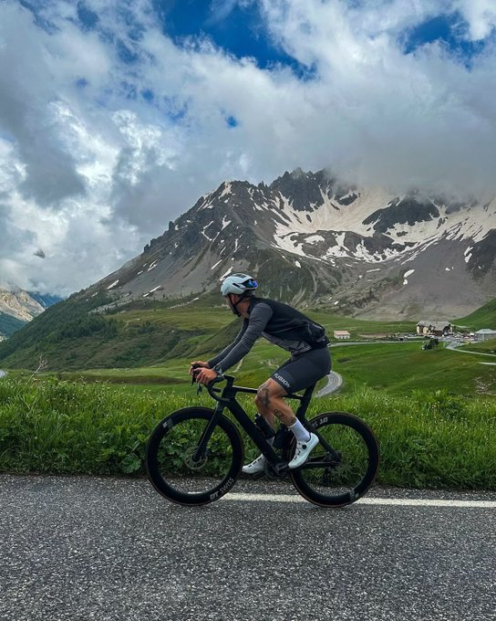 A cyclist in dark gear rides a road bike along a mountain road with snow-capped peaks and green valleys in the background.