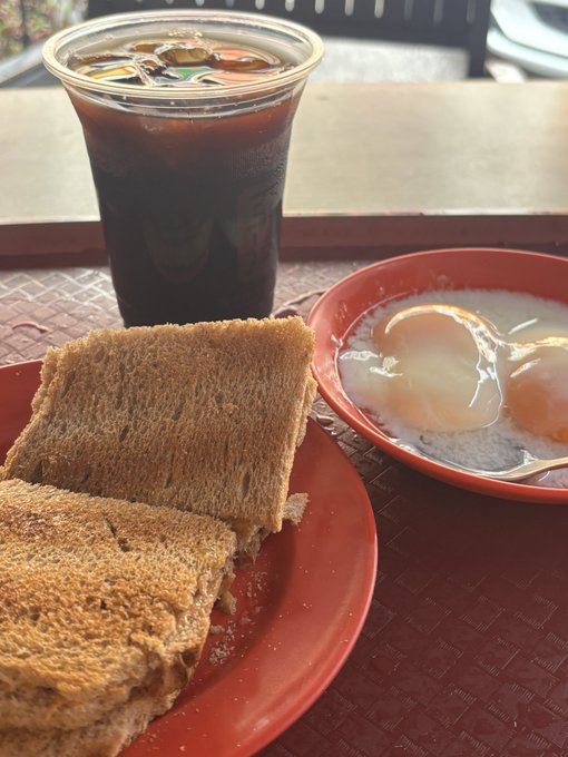 A breakfast of two slices of kaya toast, two soft-boiled eggs in a red bowl, and a cup of iced coffee on a red tray.