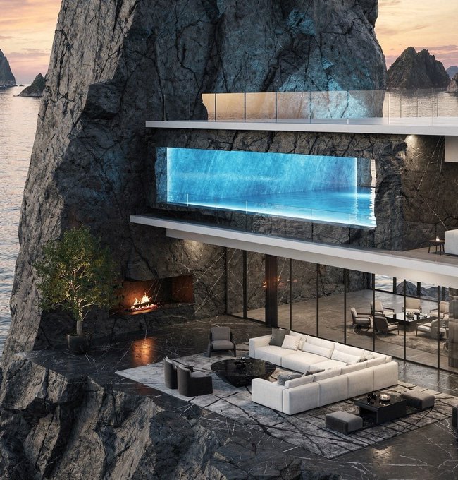 The image features an impressive modern house built into a rock, with a large glass window overlooking the ocean. The house has a white couch placed near the center of the room, surrounded by several chairs. A fireplace can be seen in one corner of the room, adding warmth and coziness to the space. The overall design and layout of the house create an interesting contrast between the natural rock formation and the man-made interior.