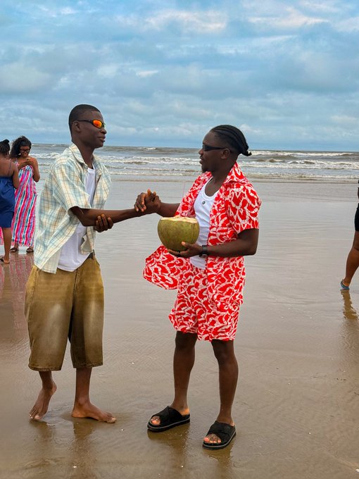 Two young men shaking hands on a beach, with one holding a coconut and wearing a red patterned shirt, while the other wears a plaid shirt and reflective sunglasses.