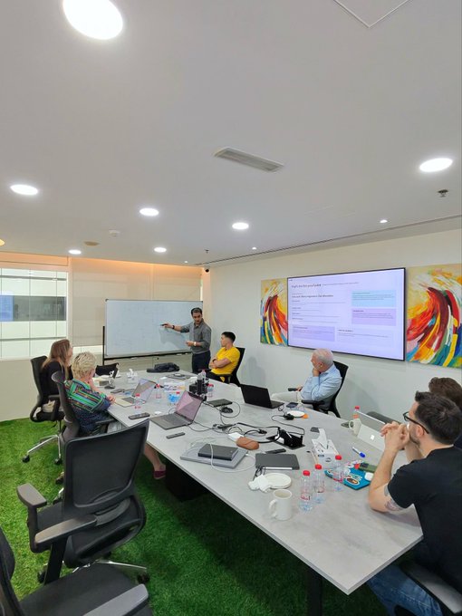 A presenter stands at a whiteboard leading a meeting with several seated participants around a conference table at $ZIG HQ, with a large screen displaying slides in the background. The whiteboard being actively used for collaboration aligns with the post highlighting its return to full use.