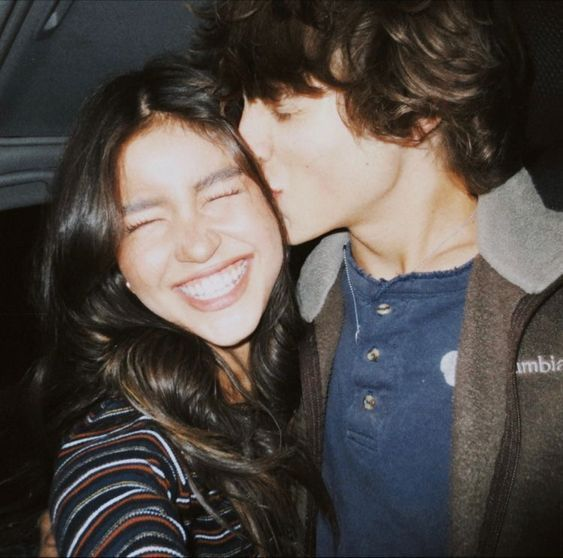 A film-style photograph captures a smiling young woman with braces being kissed on the cheek by a young man in a Columbia jacket.