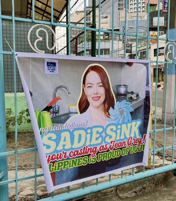 A congratulatory banner attached to a fence showing a woman's photo, with text reading 'Congratulations! SADIE SINK your casting as Jean Grey! LIPPINES is PROUD OF UUU'.