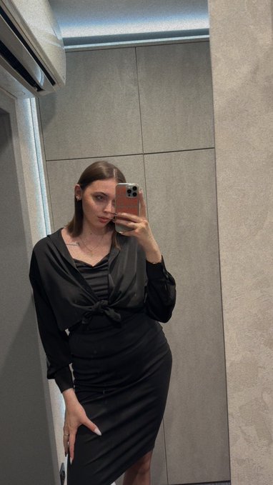 A mirror selfie of a young woman with light brown hair wearing a black long-sleeved wrap dress, standing in a room with grey tiled walls and a white air conditioning unit.