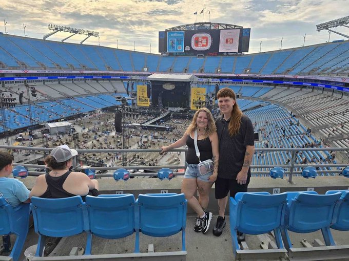 The image shows a group of people at an outdoor event, possibly a concert or sports game, standing together near some blue chairs. Two main individuals are posing for a photo - one woman with long hair and a black top, and a man wearing shorts. They appear to be enjoying themselves as they take the picture. In addition to these two main figures, there are other people in the group taking part in the event or watching from the sidelines. One person is wearing a baseball cap. The chairs are blue and scattered around the area, providing seating for attendees of the outdoor event.