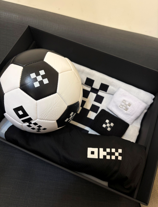 The image features a soccer ball, a black and white shirt, and a towel inside of a box. The soccer ball has a logo on it that says "OKX", which could be an indication of a brand or sponsor for the sports equipment. The black and white shirt is placed next to the soccer ball in the box, while the towel is positioned separately outside the box. The image also includes text elements such as "@mia_okx" and "@okxchinese".
