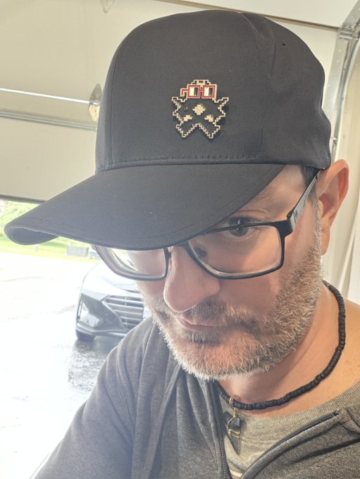 A close-up selfie of a bearded man wearing glasses, a black cap with a pixel art fox design, and a necklace, standing under a covered area.