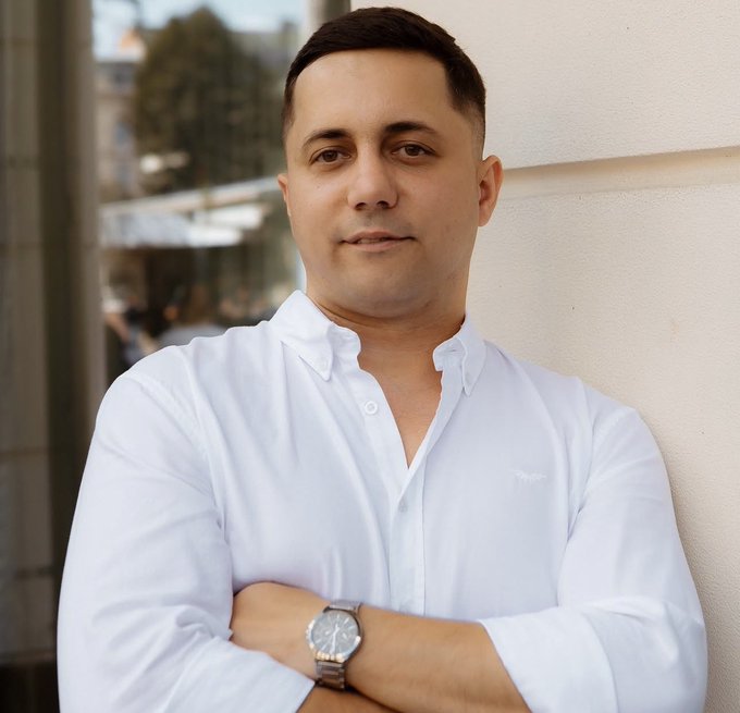 A man in a crisp white button-down shirt stands with his arms crossed, wearing a silver wristwatch, with a softly blurred outdoor architectural background.