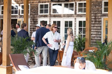 A group of people dressed in formal and semi-formal attire mingling at an outdoor social gathering near a shingled building.