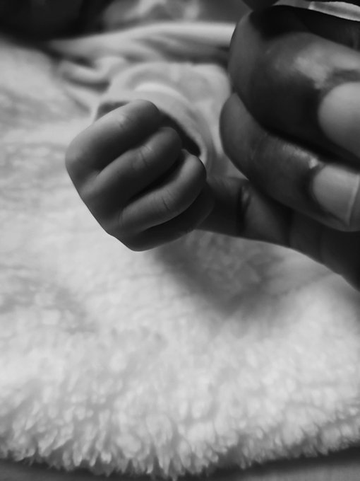 A black-and-white close-up photograph showing a tiny baby hand gripping a larger adult finger against a soft blanket.