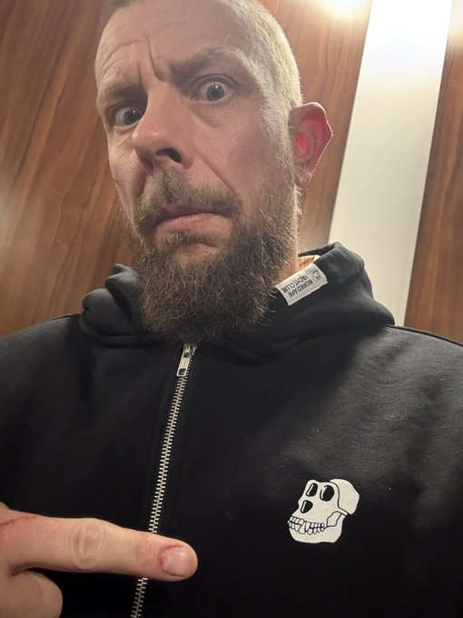 A bearded man with wide eyes points to a small white skull logo on his black hoodie, visually comparing himself to the skull associated with the Bored Ape Yacht Club NFT collection. The caption playfully references his self-identification as a "Bored Ape" by drawing attention to the matching emblem on his clothing.