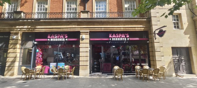 The exterior storefront of Kaspa's Desserts, featuring pink signage, outdoor seating with wicker chairs and tables, and the number 17 and 19 visible on the windows.