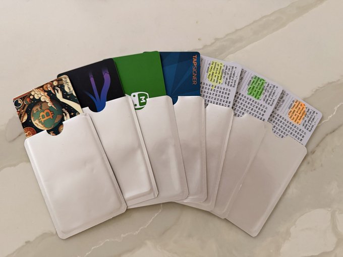 A fan of white card sleeves each partially revealing a colorful backing—including one labeled "TAPSIGNER" and another marked with a "₿" Bitcoin logo—fanned out across a marble surface, illustrating a collection of a dozen hardware signing devices. The image visually reinforces the post's enthusiasm for owning numerous Tapsigner devices for Bitcoin key security.