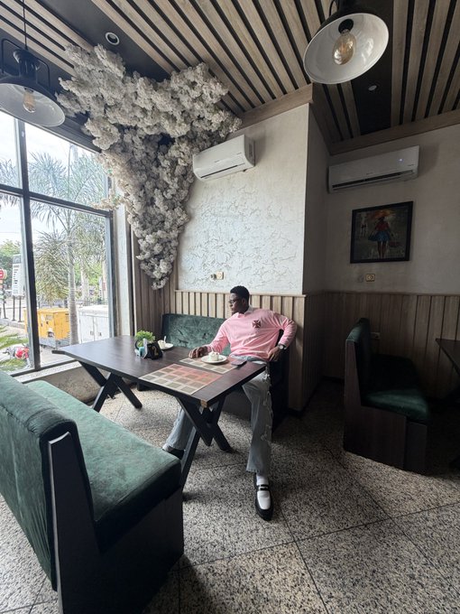 A man in a pink sweatshirt sits at a wooden table in a cafe with green velvet seating, decorative cloud-like ceiling installation, and large windows.