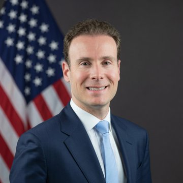 A professional headshot shows a man in a dark suit and light blue tie smiling in front of an American flag, likely depicting CFTC Chair Michael Selig who is pictured calling on Congress to pass the Clarity Act to establish regulatory standards for cryptocurrency and protect consumers.