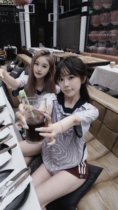 A selfie of two young women at a restaurant, one holding a glass of dark soda with a straw, with plates and cutlery visible on the table.