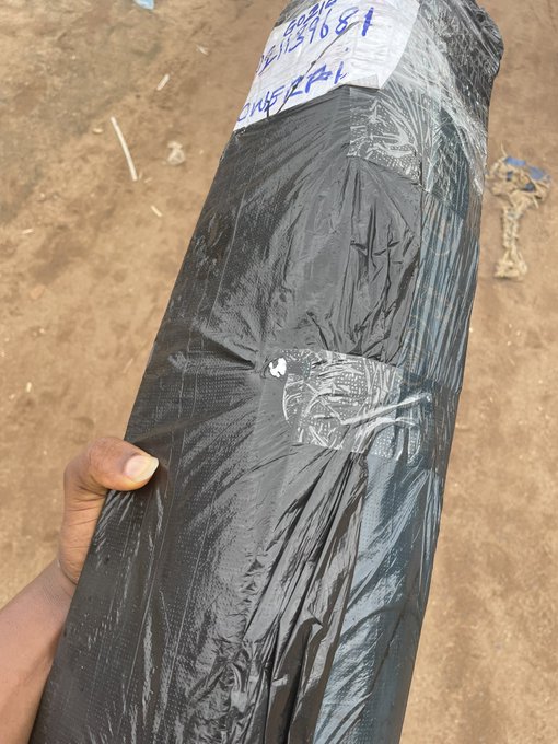 A hand holds up a long, black plastic-wrapped package on a dirt ground, with a shipping label showing handwritten numbers "904687" and "1989681." The image relates to the post by showing the "package that just arrived from Lagos," which the trader purchased using profits from $ANSEM to create upcoming content.