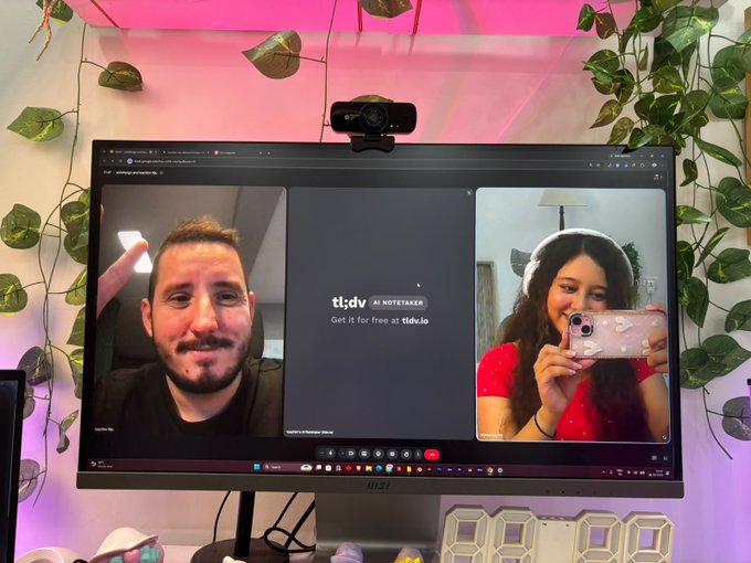 A computer monitor displays a video call between a man in a black shirt and a young woman in a red top holding up a phone, with the 'tl;dv AI NOTETAKER' logo and 'Get it for free at tl;dv.io' visible on screen.