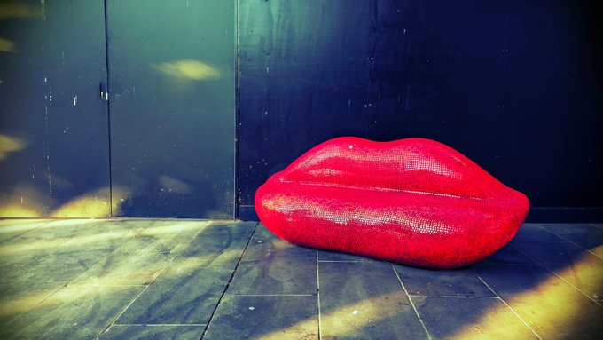 A sparkling red sequined lip-shaped sculpture sits on a stone-tiled floor against a dark backdrop, embodying the whimsical, surreal nature of artistic expression—as if conjured by magic. The contrast of the bold, glittering lips with the moody, sun-dappled setting evokes the transformative power of art to turn ordinary spaces into something enchanting.