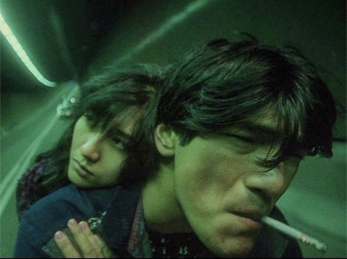 A moody, green-tinted cinematic photo of a young man and woman, with the man smoking a cigarette in the foreground.