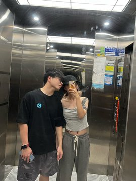 The image shows a man and woman standing together inside an elevator, taking a selfie with their cell phones. The couple appears to be enjoying themselves as they capture this moment. The background of the photo features a metal wall that adds a modern touch to the scene.