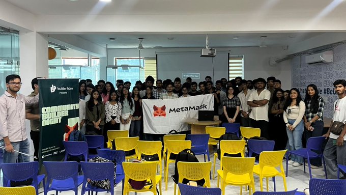 A group photo of participants at a BuilderSsoi Presents event, with attendees holding a MetaMask banner and wearing 'COMMUNITY BUILDER NIGHT' lanyards in a room with colorful chairs.