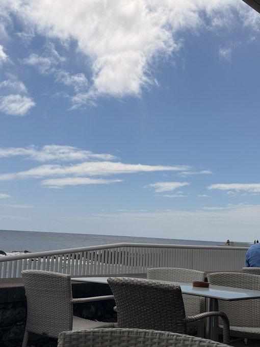 A scenic oceanfront patio with white wicker chairs and tables under a bright, partly cloudy sky — the kind of serene, trustworthy backdrop someone could share before requesting a Solana wallet address, though real crypto scams typically impersonate others and this image doesn't verify any legitimate transaction.
