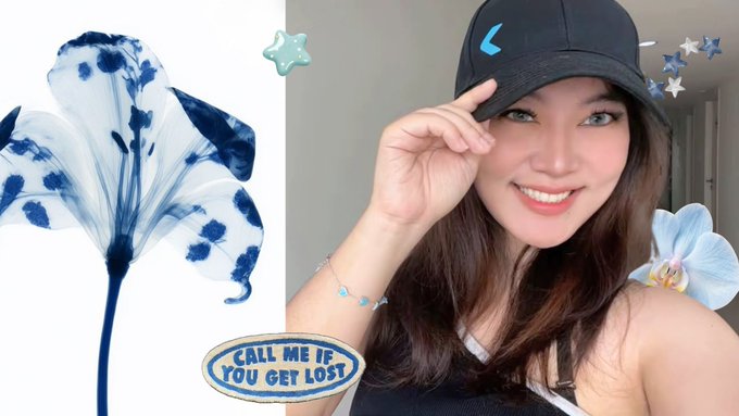 A young woman smiles while tipping a navy baseball cap adorned with a blue Typhoon logo, styled with a delicate bracelet, alongside an indigo tie-dyed flower and a "CALL ME IF YOU GET LOST" patch — merchandise that complements her relaxed, trendy aesthetic.