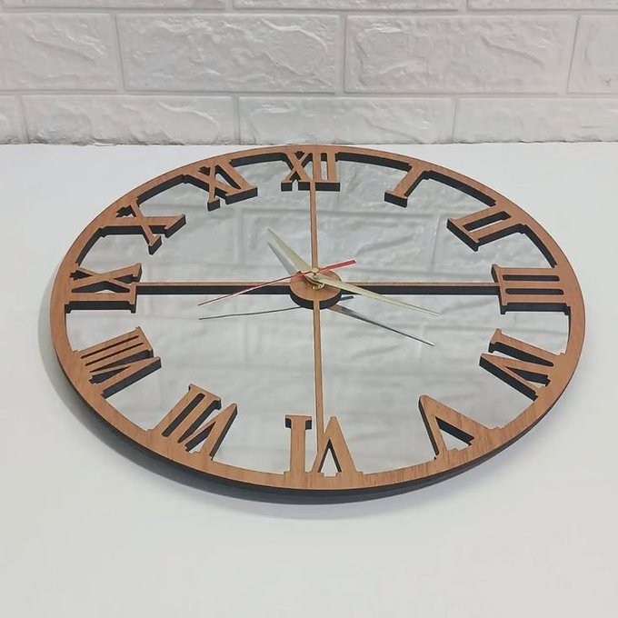 A large minimalist wooden wall clock with Roman numerals and silver hands sits against a white brick wall on a white surface.