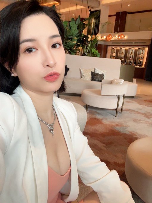 A woman with short dark hair wearing a white blazer and coral top takes a selfie in an elegant lobby setting, accessorized with a sparkling necklace.